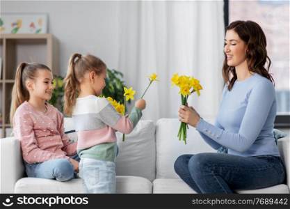 people, family and holidays concept - two daughters giving daffodil flowers to happy mother at home. daughters giving daffodil flowers to happy mother