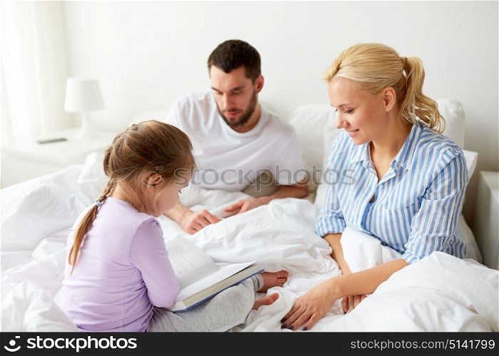 people, family and morning concept - happy child with parents reading book in bed at home. happy family reading book in bed at home
