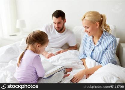 people, family and morning concept - happy child with parents reading book in bed at home. happy family reading book in bed at home