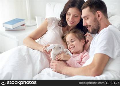 people, family and morning concept - happy child with toy rabbit and parents in bed at home. happy child with toy and parents in bed at home