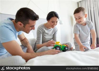people, family and morning concept - happy child with toy tractor and parents playing in bed at home or hotel room. happy family in bed at home or hotel room