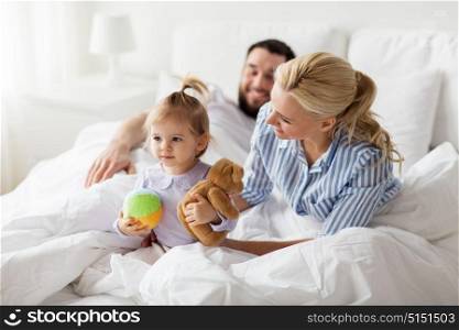 people, family and morning concept - happy child with toys and parents in bed at home. happy child with toys and parents in bed at home