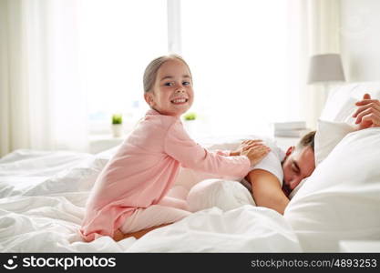 people, family and morning concept - happy little girl waking her sleeping father up in bed at home