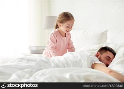 people, family and morning concept - happy little girl waking her sleeping father up in bed at home
