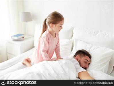 people, family and morning concept - happy little girl waking her sleeping father up in bed at home. little girl waking her sleeping father up in bed