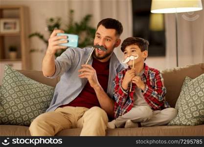 people, family and technology concept - happy father and little son with mustaches party props taking selfie by smartphone sitting on sofa at home in evening. father and son taking selfie at home