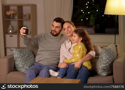 people, family and technology concept - happy father, mother and little daughter taking selfie by smartphone at home at night. happy family taking selfie at home at night