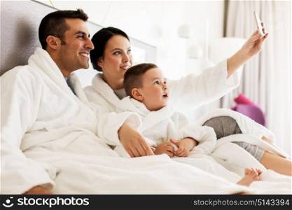 people, family and technology concept - happy mother, father and little boy in bathrobe taking selfie by smartphone in bed or hotel room. happy family with smartphone in bed at hotel room
