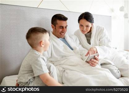 people, family and technology concept - happy mother, father and little boy with smartphone in bed at hotel room. happy family with smartphone in bed at hotel room