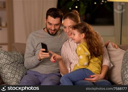 people, family and technology concept - happy mother, father and little daughter with smartphone at home at night. happy family with smartphone at home at night