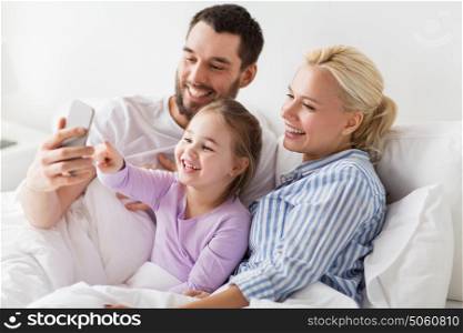 people, family and technology concept - happy mother, father and little girl taking selfie with smartphone in bed at home. happy family taking selfie by smartphone at home