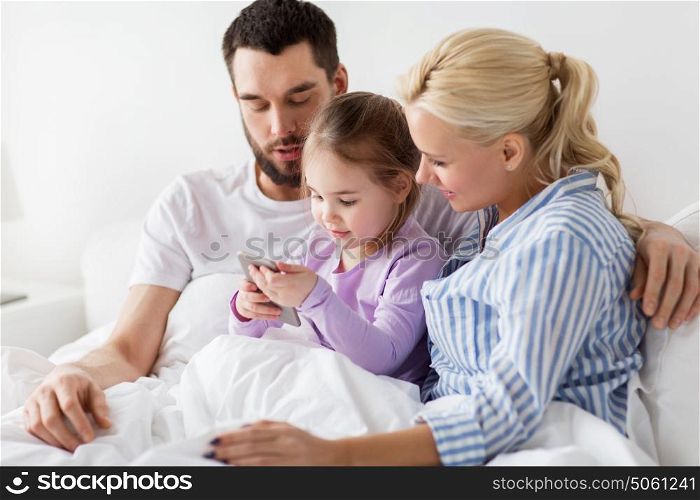 people, family and technology concept - happy mother, father and little girl with smartphone in bed at home. happy family with smartphone in bed at home