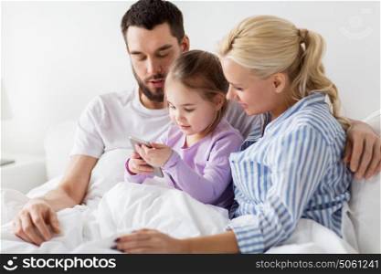 people, family and technology concept - happy mother, father and little girl with smartphone in bed at home. happy family with smartphone in bed at home