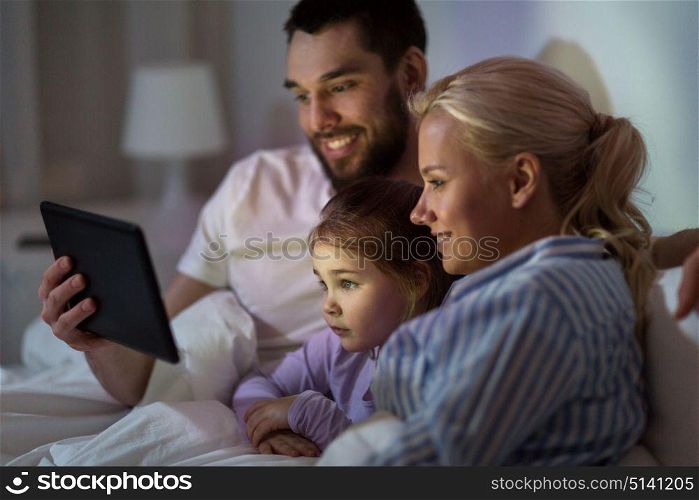 people, family and technology concept - happy mother, father and little girl with tablet pc computer in bed at night home. happy family with tablet pc in bed at home