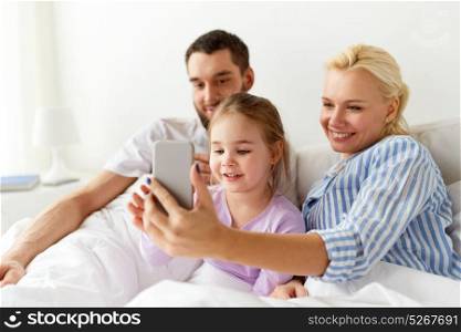 people, family and technology concept - happy mother, father and little girl with smartphone in bed at home. happy family with smartphone at home