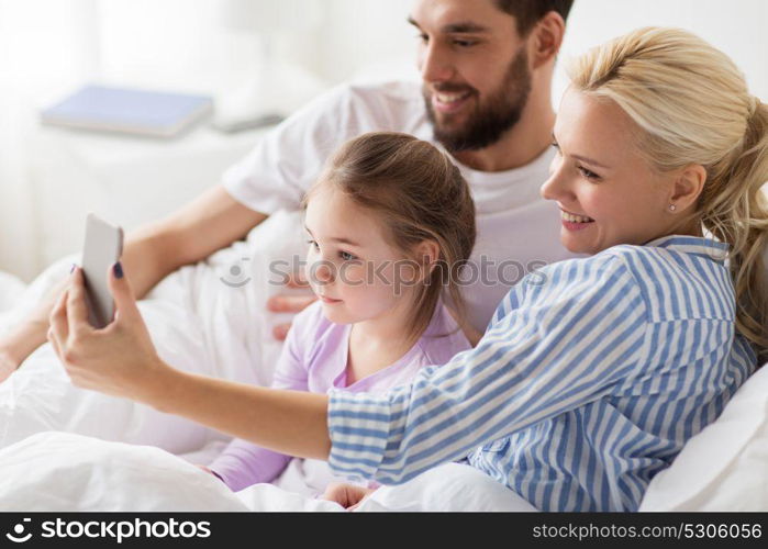 people, family and technology concept - happy mother, father and little girl taking selfie with smartphone in bed at home. happy family taking selfie by smartphone at home