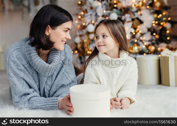 People, family, celebration and holidays concept. Mother and daughter with attractive appearance look at each other`s eyes, lie on warm white carpet near decorated New Year tree and gift box