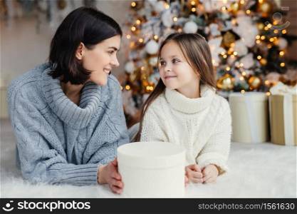 People, family, celebration and holidays concept. Mother and daughter with attractive appearance look at each other`s eyes, lie on warm white carpet near decorated New Year tree and gift box