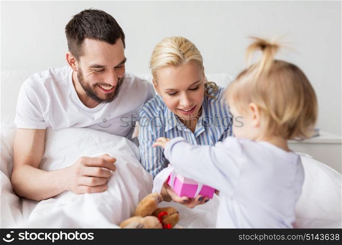 people, family, holidays and morning concept - happy little girl and parents with gift box in bed at home. happy family with gift box in bed at home