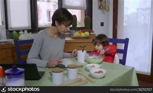 People, family life, fun, lifestyle, child rearing, motherhood. Busy woman talking on the phone in kitchen at home, multitasking mom cooking, working, smiling with female child, baby girl, daughter