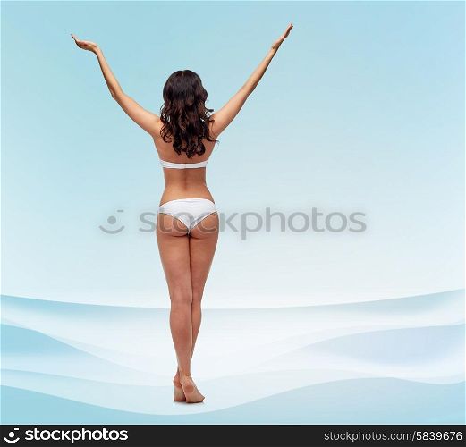 people, fashion, beauty, summer and beach concept - young woman posing in white bikini swimsuit with raised hands from back over blue waves background