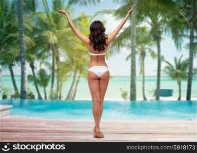 people, fashion, beauty, summer and travel concept - young woman posing in white bikini swimsuit with raised hands from back over tropical beach with palm trees and pool at hotel resort background