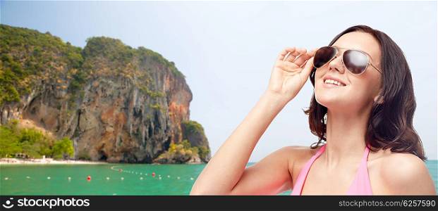 people, fashion, summer accessories and beach concept - happy young woman in sunglasses and pink swimsuit looking up over sea and rock background