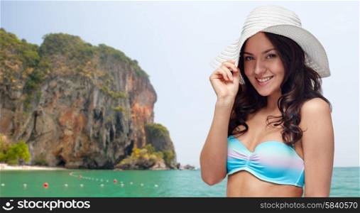 people, fashion, summer and beach concept - happy young woman in bikini swimsuit and sun hat over sea and rock background