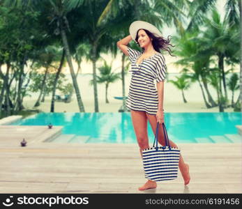 people, fashion, summer and beach concept - happy young woman in summer clothes and sun hat with bag over swimming pool at beach resort
