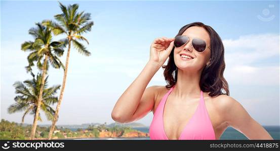 people, fashion, summer and travel concept - happy young woman in sunglasses and pink bikini swimsuit over Sri Lanka beach with palms background