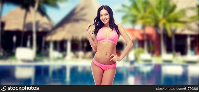 people, fashion, summer vacation and travel concept - happy sexy young woman posing in pink bikini swimsuit over hotel resort with swimming pool, bungalow and palm trees background