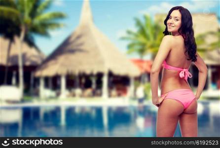 people, fashion, summer vacation and travel concept - happy sexy young woman in pink bikini swimsuit looking back over hotel resort with swimming pool, bungalow and palm trees background