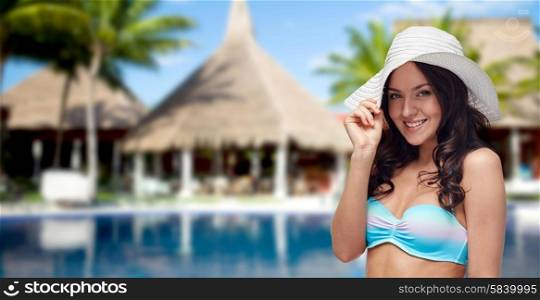 people, fashion, summer vacation and travel concept - happy young woman in bikini swimsuit and sun hat over swimming pool, bungalow and palm trees at hotel resort background