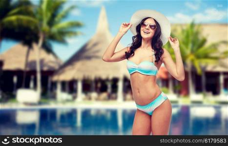 people, fashion, summer vacation and travel concept - happy young woman in bikini swimsuit, sunglasses and sun hat over swimming pool, bungalow and palm trees at hotel resort background