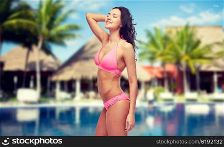 people, fashion, summer vacation and travel concept - happy young woman posing in pink bikini swimsuit over hotel resort with swimming pool, bungalow and palm trees background