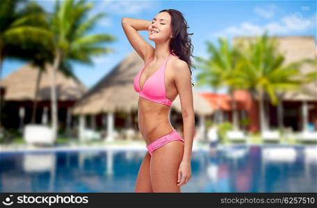 people, fashion, summer vacation and travel concept - happy young woman posing in pink bikini swimsuit over hotel resort with swimming pool, bungalow and palm trees background