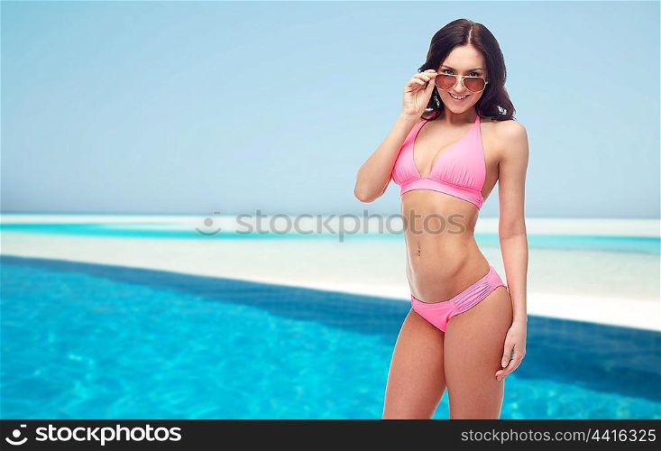 people, fashion, swimwear, summer and beach concept - happy young woman in sunglasses and pink swimsuit looking at you over maldives beach with swimming pool background
