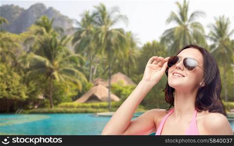 people, fashion, swimwear, summer and beach concept - happy young woman in sunglasses and pink swimsuit looking up over swimming pools and palm trees background