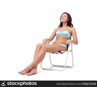 people, fashion, swimwear, summer and beach concept - happy young woman in bikini swimsuit sunbathing on folding chair