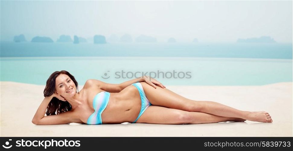 people, fashion, swimwear, summer and beach concept - happy young woman lying in bikini swimsuit over infinity edge pool at hotel resort background