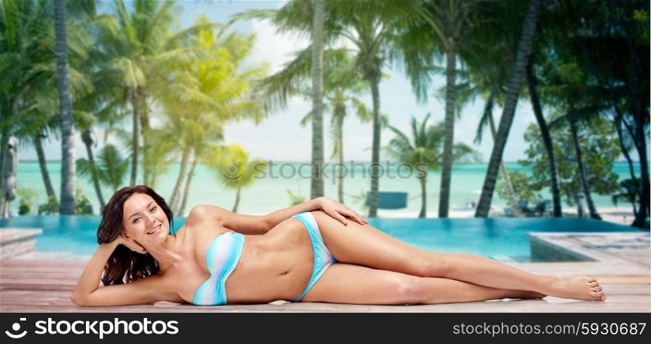 people, fashion, swimwear, summer and beach concept - happy young woman lying in bikini swimsuit over tropical beach with palm trees and pool at hotel resort background