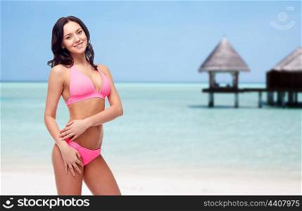 people, fashion, swimwear, summer and beach concept - happy young woman posing in pink bikini swimsuit over exotic tropical beach with bungalow in sea background