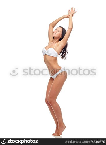 people, fashion, swimwear, summer and beach concept - happy young woman posing in white bikini swimsuit dancing with raised hands