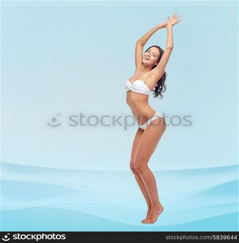 people, fashion, swimwear, summer and beach concept - happy young woman posing in white bikini swimsuit dancing with raised hands over blue waves background