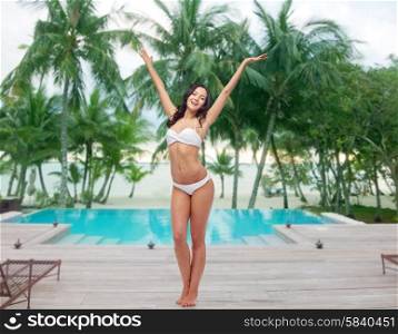 people, fashion, swimwear, summer and beach concept - happy young woman posing in white bikini swimsuit dancing with raised hands over swimming pool at beach resort