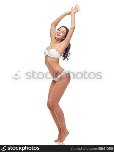 people, fashion, swimwear, summer and beach concept - happy young woman posing in white bikini swimsuit dancing with raised hands