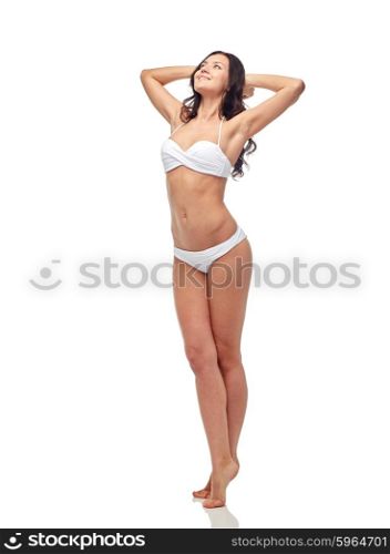 people, fashion, swimwear, summer and beach concept - happy young woman posing in white bikini swimsuit with raised hands looking up