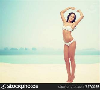 people, fashion, swimwear, summer and beach concept - happy young woman posing in white bikini swimsuit with raised hands over infinity pool at beach resort