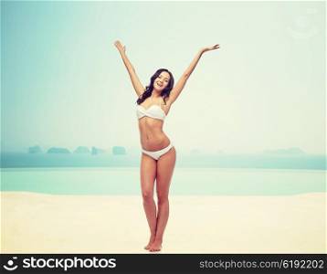 people, fashion, swimwear, summer and beach concept - happy young woman posing in white bikini swimsuit dancing with raised hands over infinity pool at beach resort
