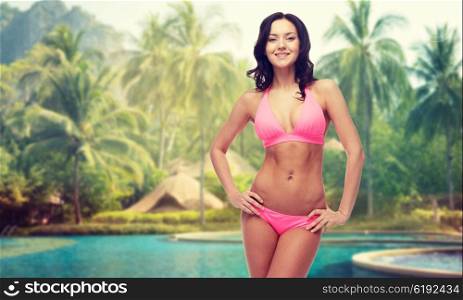 people, fashion, swimwear, summer and beach concept - happy young woman posing in pink bikini swimsuit over swimming pools and palm trees background